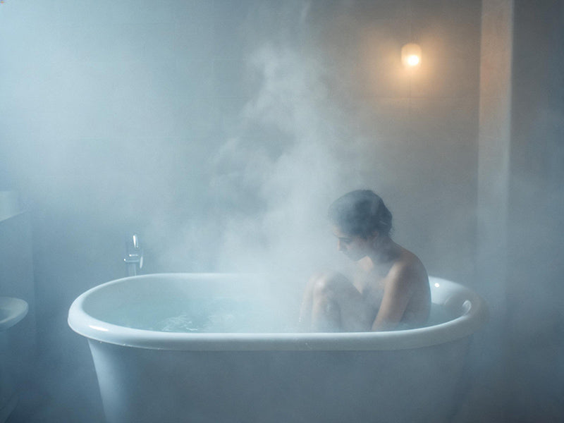 A misty bathroom scene with a clawfoot bathtub, warm steam rising, and soft, dim light filtering through the fog, capturing the intimacy and vulnerability of listening to Raveena's "Temptation"